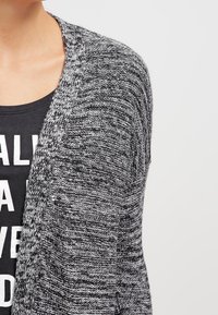 Szary i czarny dziergany kardigan z otwartym przodem, z teksturowanym wzorem i luźnym krojem, noszony na czarnej bluzce z białym napisem.