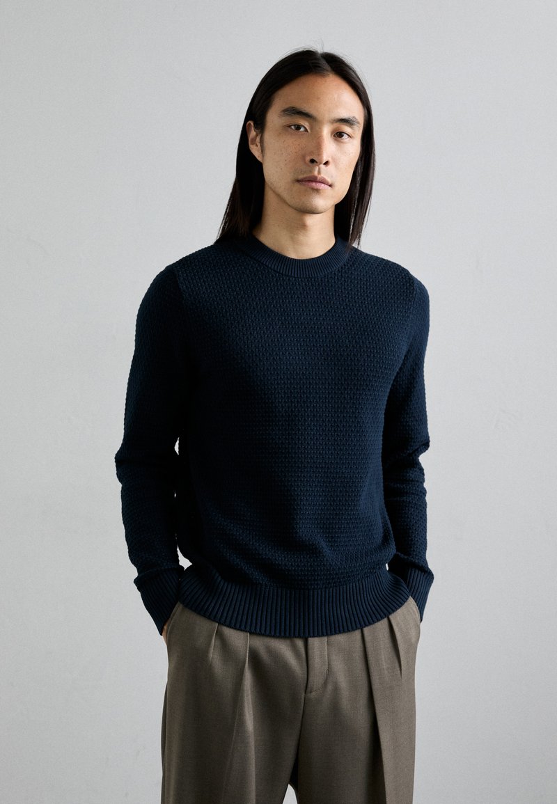 Marineblauer Strickpullover aus strukturiertem Stoff, mit geripptem Kragen und Bündchen, kombiniert mit hellgrauen, gefalteten Hosen. Einfaches, modernes Design.