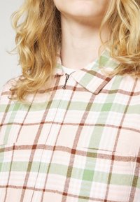 Pink and green plaid shirt with a collar, featuring brown accents and a zip closure. The fabric appears soft with a textured finish.