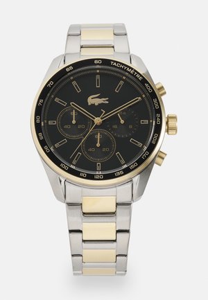 Reloj cronógrafo redondo con esfera negra, acentos dorados, correa de acero inoxidable plateada y escala tacómetro; presenta tres subesferas.