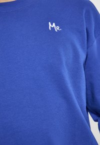Blauwe katoenen T-shirt met een ronde halslijn; voorzien van witte geborduurde tekst "Mr." bij de borst; heeft een gladde textuur.