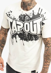 Majica od pamuka boje kreme s velikim crnim logotipom "TAPOUT" i apstraktnim splatterskim dizajnom. Ima kratke rukave i okrugli izrez.