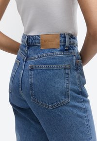 Nærbillede af en person iført højtsiddende blå cowboybukser med en brun "WEEKDAY" læderlappe og en hvid ribbet tanktop.