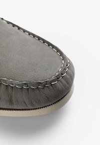 Grå læder moccasin sko med en afrundet tå, der har kontrasterende hvid syning og en struktureret, blød overflade. Gummisålen er synlig.