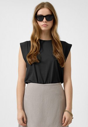 Jeune femme aux cheveux longs et ondulés, portant un haut noir sans manches, une jupe beige, des lunettes de soleil noires en forme d'œil de chat et un bracelet doré, debout devant un fond blanc.