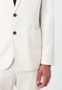 Lichtkleurige blazer van gladde stof, met twee bruine knopen, een opgestikte zak en een getailleerde pasvorm.