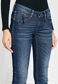 Dark blue denim jeans with a slim fit, featuring front pockets and a zippered coin pocket with a black zipper pull. Visible stitching details.