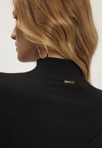 Czarna, prążkowana tkanina odzieżowa z wysokim kołnierzem. Posiada złotą tabliczkę z logo z tyłu oraz modelkę noszącą duże złote kolczyki w kształcie koła.