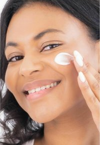 En hudvårdsprodukt appliceras på kinden; krämig vit konsistens, slät finish, modellen har en naturlig lyster i huden, ljus bakgrund.