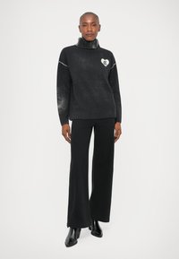 Sweater preto de gola alta com um logótipo de coração branco, textura suave e gola canelada. Combinado com calças largas pretas e botas de salto.