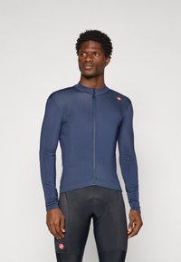 Homme portant un maillot de cyclisme ajusté à manches longues bleu marine et un short de cyclisme rembourré noir, se tenant devant un fond uni.