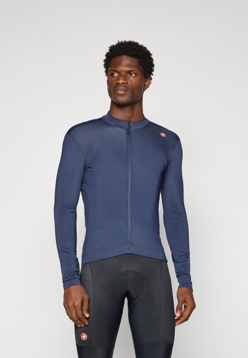 Homme portant un maillot de cyclisme ajusté à manches longues bleu marine et un short de cyclisme rembourré noir, se tenant devant un fond uni.