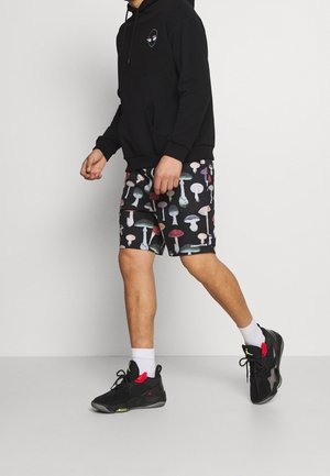 Schwarzer Hoodie mit Fronttasche; gemusterte Shorts mit bunten Pilzen auf schwarzem Hintergrund; schwarze Sportschuhe mit roten Akzenten.