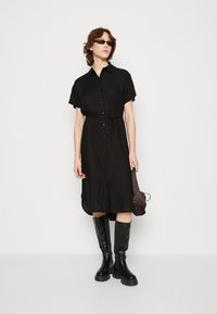 Vestito nero a camicia con maniche corte, chiusura a bottoni frontale, cintura in vita e spacchi laterali; abbinato a stivali neri alti fino al ginocchio e una borsa scura.