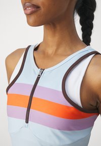 Haut de sport bleu clair avec des rayures orange, violet et marron. Présente une fermeture éclair à l'avant et un liseré marron contrastant le long de l'encolure.