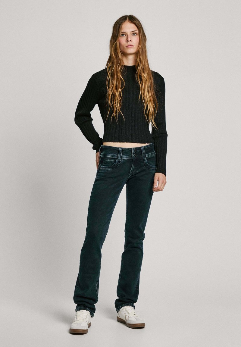 Dunkelblaue Denim-Jeans mit leichtem Schlag, kombiniert mit einem gerippten, schwarzen, kurzen Pullover. Weiße Sneakers mit braunen Akzenten runden den Look ab.