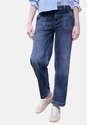 Persoon die donkerblauwe jeans met ruime pasvorm en grote voorzakken draagt, lichtblauw overhemd en beige sneakers met witte veters.