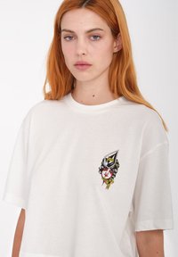 Camiseta blanca de algodón con mangas cortas, que presenta un gráfico bordado de la cara de una mujer con un tocado decorativo y una bufanda.