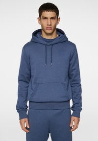 Blå huvtröja med en framficka av typ kangaroo, dragsko i huvan, ribbade ärmslut och midja. Har en liten logotyp på bröstet.