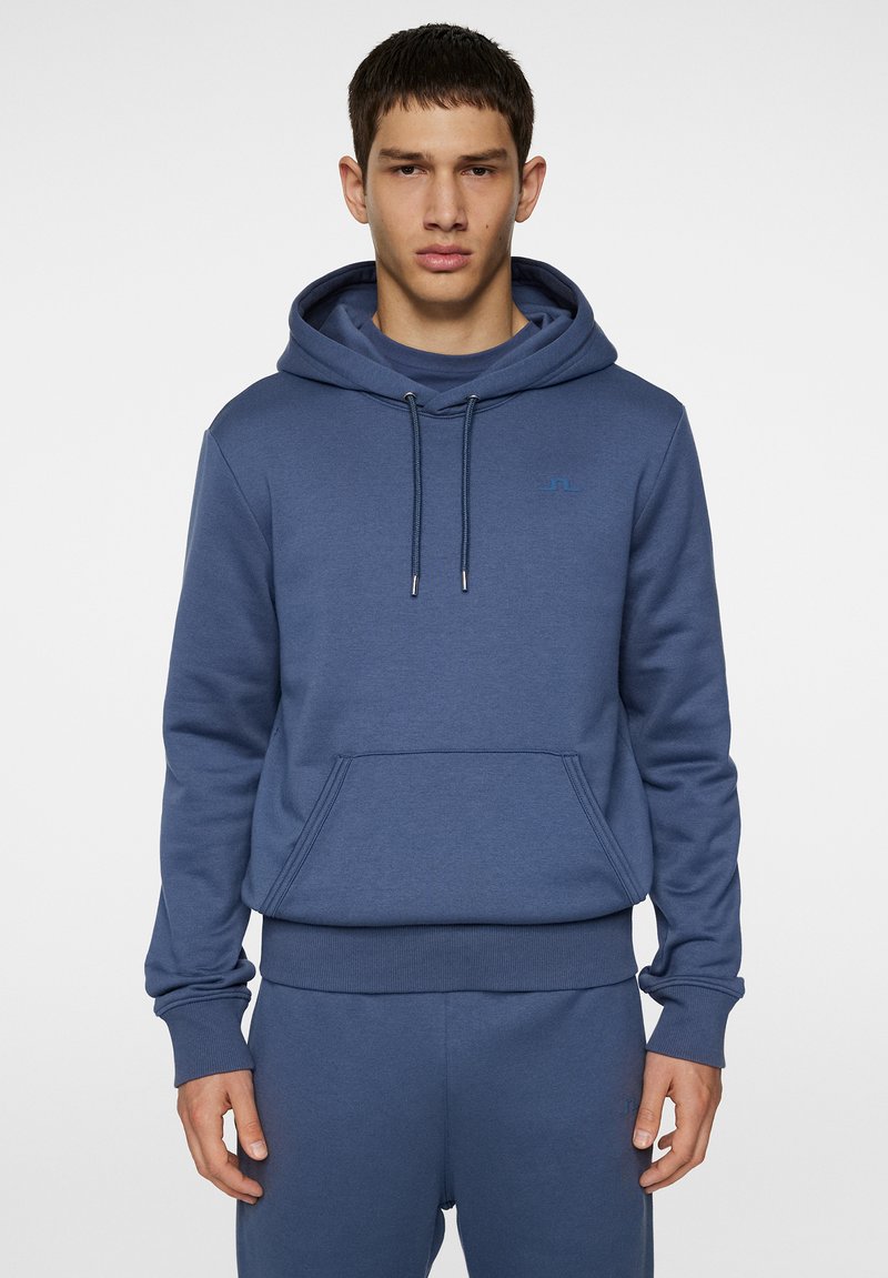 Blå huvtröja med en framficka av typ kangaroo, dragsko i huvan, ribbade ärmslut och midja. Har en liten logotyp på bröstet.