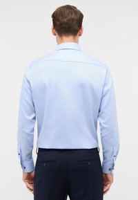 Camicetta azzurra chiara a maniche lunghe con colletto classico, dettagli con bottoni sui polsini e una texture liscia, abbinata a pantaloni scuri.