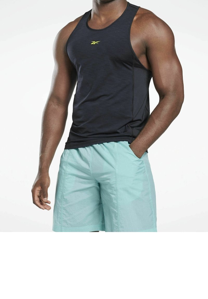 Torse masculin portant un débardeur de sport noir et un short de sport bleu clair, une main dans la poche, sur un fond blanc uni.