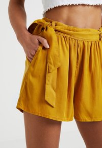 Żółte szorty o wysokim stanie z lekkiego materiału, z zawiązywanym pasem, bocznymi kieszeniami oraz plisowanym wzorem.