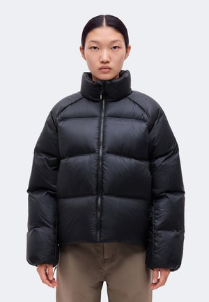 Un modèle est de face, vêtu d’une doudoune noire matelassée à col montant et d’un pantalon marron, sur un fond clair et uni.