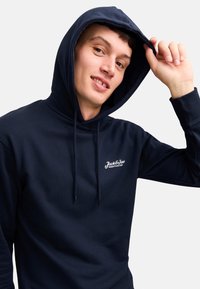 Granatowy hoodie z miękkiego materiału, z regulowanym kapturem i małym białym logo na klatce piersiowej. Długie rękawy z dopasowanymi mankietami.