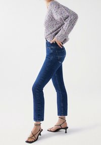 Jeans en denim bleu à taille haute, coupe slim et minimalement délavés, associés à un pull en tricot texturé de couleur lilas clair et des talons noirs à lanières.