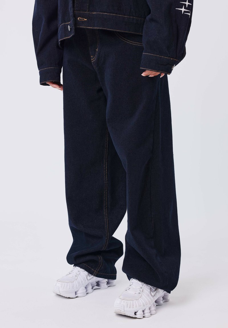 Pantalones de mezclilla de corte completo en un azul oscuro, con costuras laterales prominentes y costura dorada, combinados con zapatillas deportivas blancas y acolchadas.