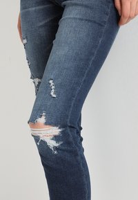 Mörkblå skinny jeans med slitna detaljer, inklusive knäskärningar och fransade kanter, tillverkade av denimtyg med en slät textur.