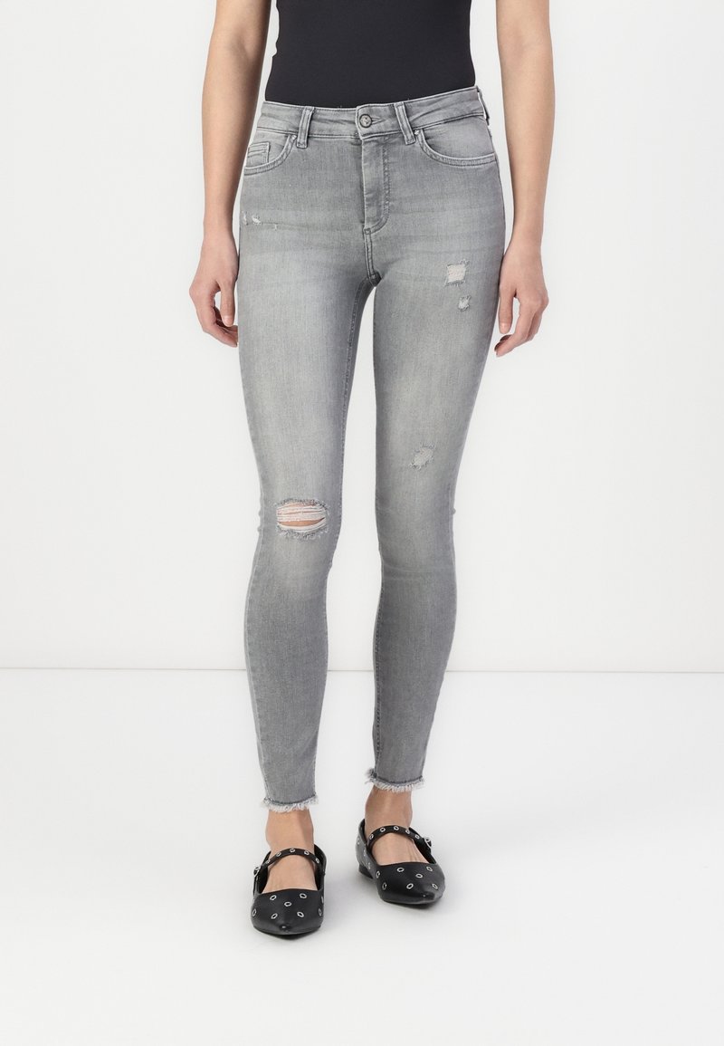 Jeans ajustados grises con detalles desgastados, que presentan desgarros en las rodillas y dobladillos deshilachados. Combinados con zapatos planos negros con remaches.