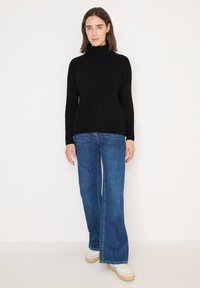 Pull noir à col roulé en maille avec texture côtelée et motif losange ; associé à un jean en denim bleu évasé et des baskets blanches.