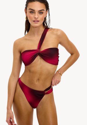 Femme portant un bikini rouge à une épaule, avec des boucles d'oreilles anneaux dorés et un bracelet en or, sur un fond blanc.