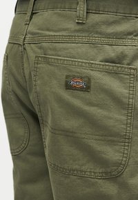 Φανελένιο παντελόνι σε ελιά χρώμα Dickies με πίσω τσέπη που φέρει ραμμένη ετικέτα με το εμπορικό σήμα σε κόκκινους, κίτρινους και μπλε χρωματισμούς.