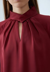 Burgundy blouse with a high collar, featuring a front keyhole and pleated detailing, made from smooth, lightweight fabric.