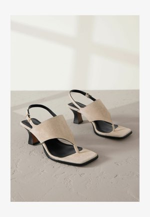 Beżowe zamszowe sandały z designem na palce, kwadratowym noskiem i blokowym obcasem. Posiadają czarny pasek w kostkę z metalowym detalem z klamrą.