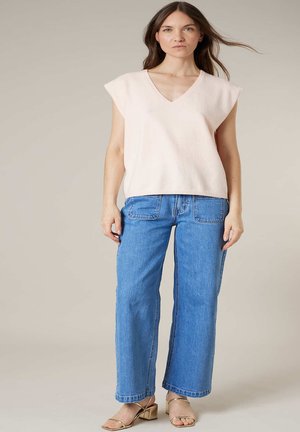 Femme portant un haut beige sans manches à col en V, un jean droit bleu avec poches avant, et des sandales dorées à bouts ouverts, debout devant un fond uni.