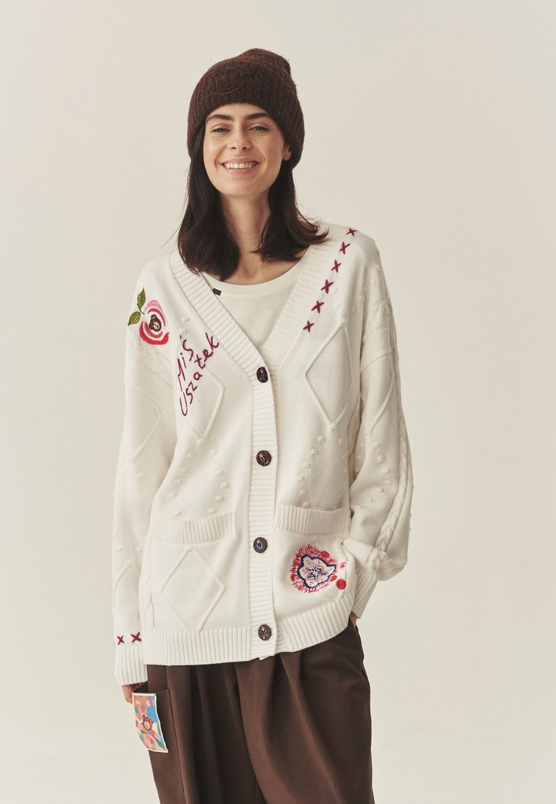 Biały dziergany sweter z fakturowanymi wzorami, haftowanymi kwiatami, dużymi kieszeniami i ciemnymi guzikami, noszony na kremowej bluzce z brązowymi spodniami.