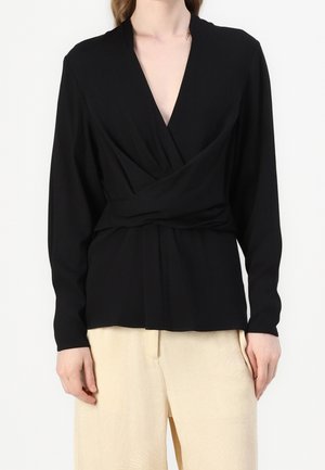 Kvinde iført en sort langærmet omslagsbluse med dyb V-hals og beige bukser, vist fra halsen til hoften.