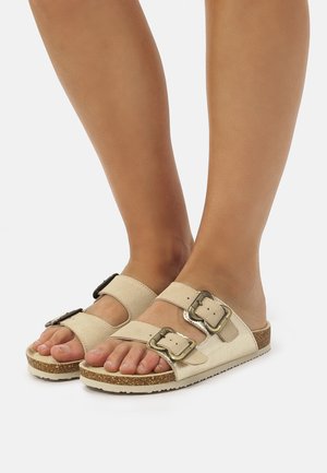 Chanclas deslizantes beige hechas de una tela suave, con dos correas ajustables que presentan hebillas metálicas y una suela de corcho texturizada.