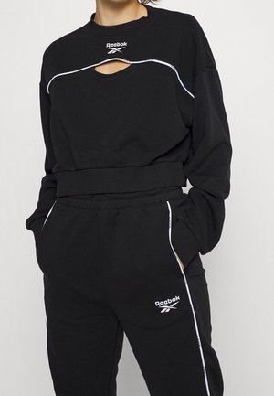 Osoba w czarnej bluzie Reebok z wycięciem na klatce piersiowej oraz dopasowanych spodniach dresowych z białym obszyciem i logo, z rękami w kieszeniach.