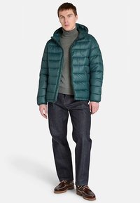 Giacca imbottita turchese con cappuccio, cuciture orizzontali, chiusura con zip, indossata sopra un dolcevita grigio e jeans scuri.