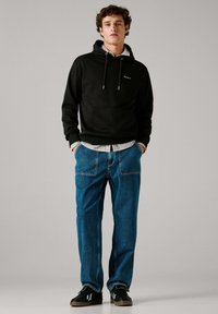 Must kapuutsiga dressipluus esitaskuga, kombineeritud lahtiste siniste teksapükstega. Mustad tossud valgete detailidega lõpetavad komplekti. Hall taust.