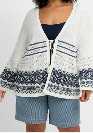Femme portant un cardigan blanc ouvert avec des motifs géométriques bleu marine sur un haut bleu marine et un short à fines rayures bleues.
