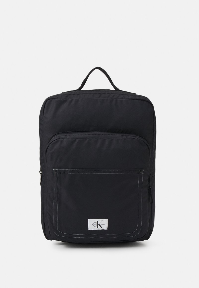 Sac à dos carré noir avec poche avant zippée et patch logo Calvin Klein, poignée supérieure unique, et texture de tissu lisse.