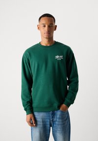 Groene sweatshirt met geribbelde manchet en halslijn, met een wit logo en tekst op het linkerkant van de borst. Draagt met blauwe jeans.