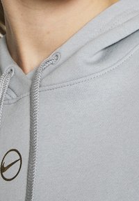 Ljusgrå huvtröja med mjuk textur, utrustad med snörning vid halsen och en liten, mörkbrun Nike-logotyp på bröstet.