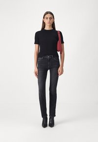Svart tettsittende t-skjorte, mørkeblå jeans og svarte ankelstøvler. En rød justerbar reim henger fra skulderen. Nøytral bakgrunn.